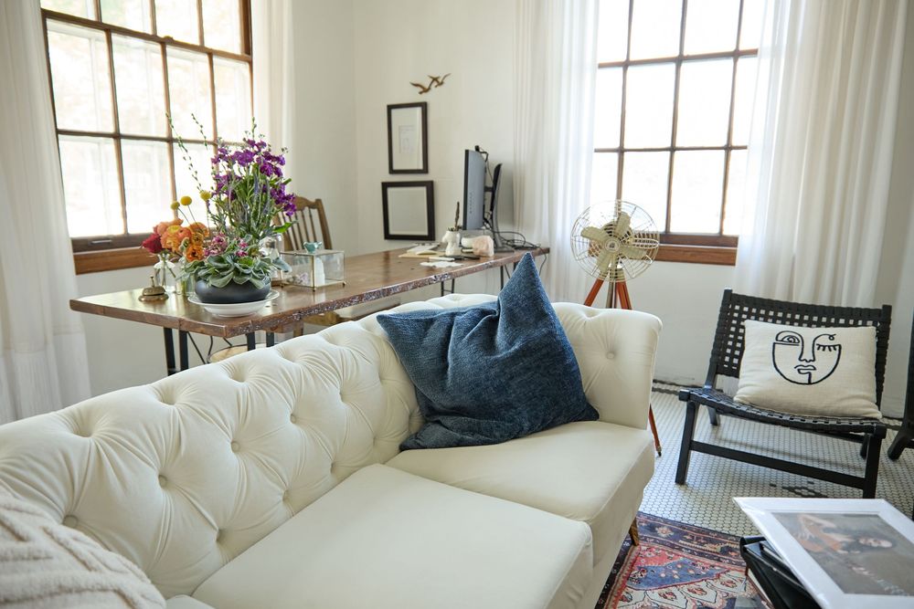 White tufted sofa with blue pillows beside large windows and gallery wall.
