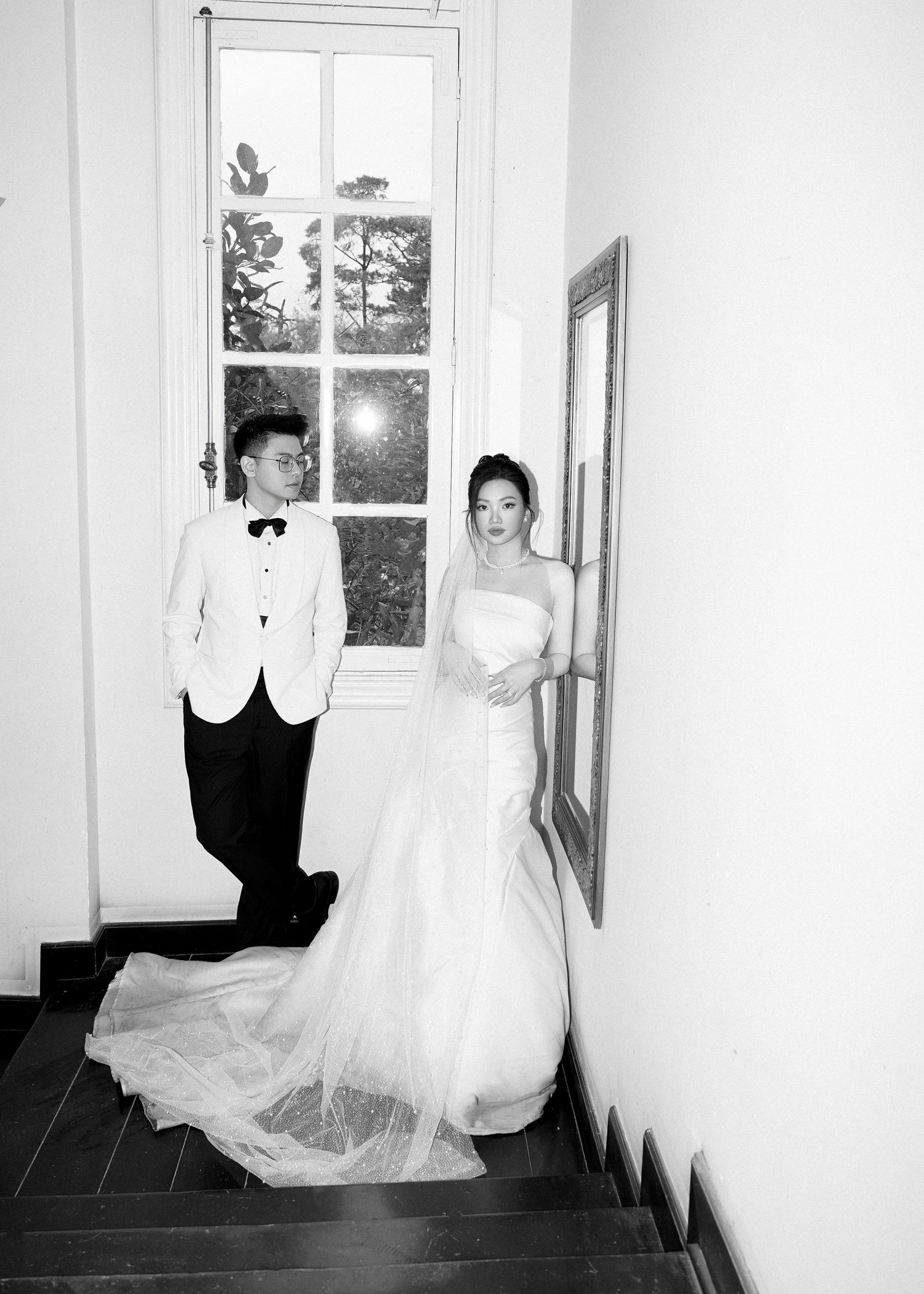Bride and groom pose elegantly on a staircase by a tall window in a black and white portrait.