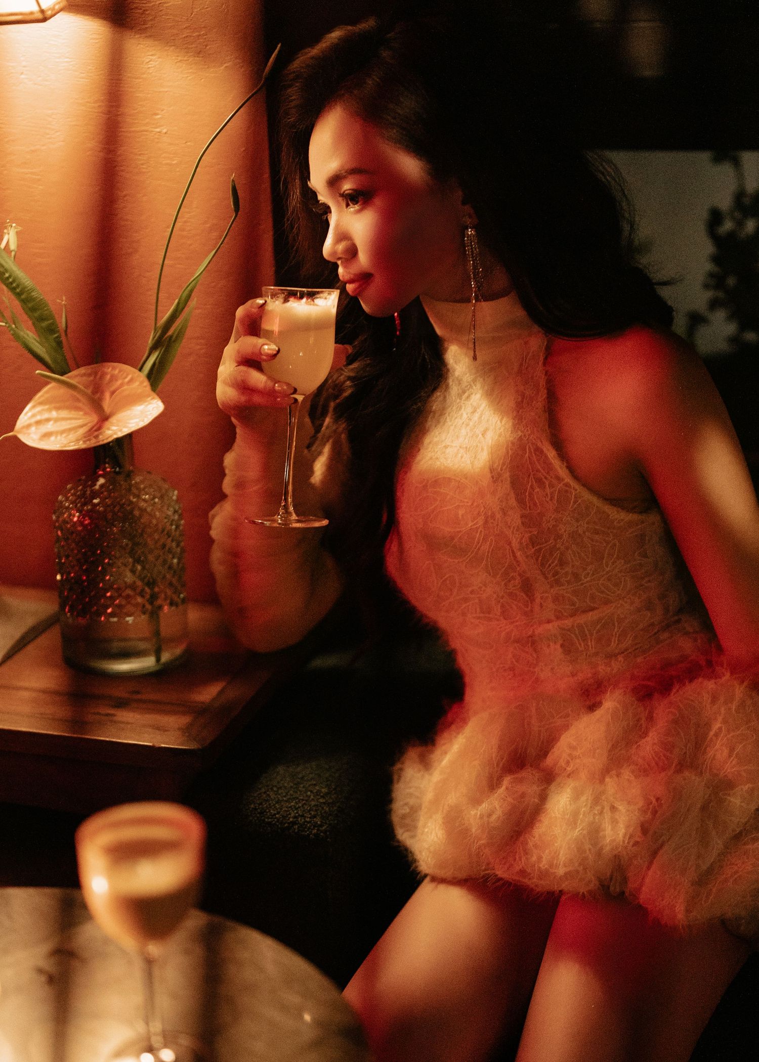 Elegant woman in white feathered dress sipping a cocktail in a warmly lit intimate bar setting.