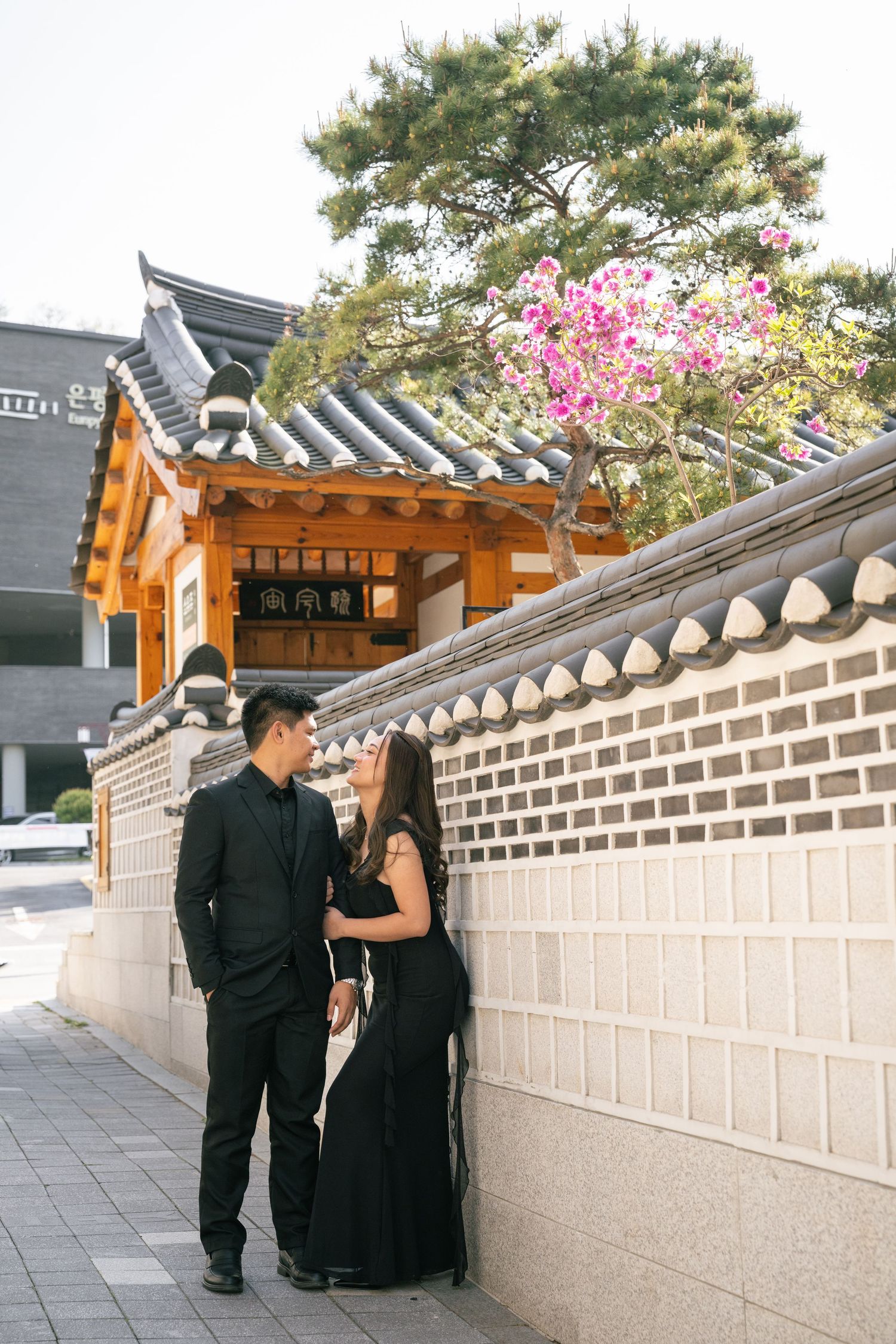 Two people in formal black attire walking along a traditional Korean stone wall with cherry blossoms visible.