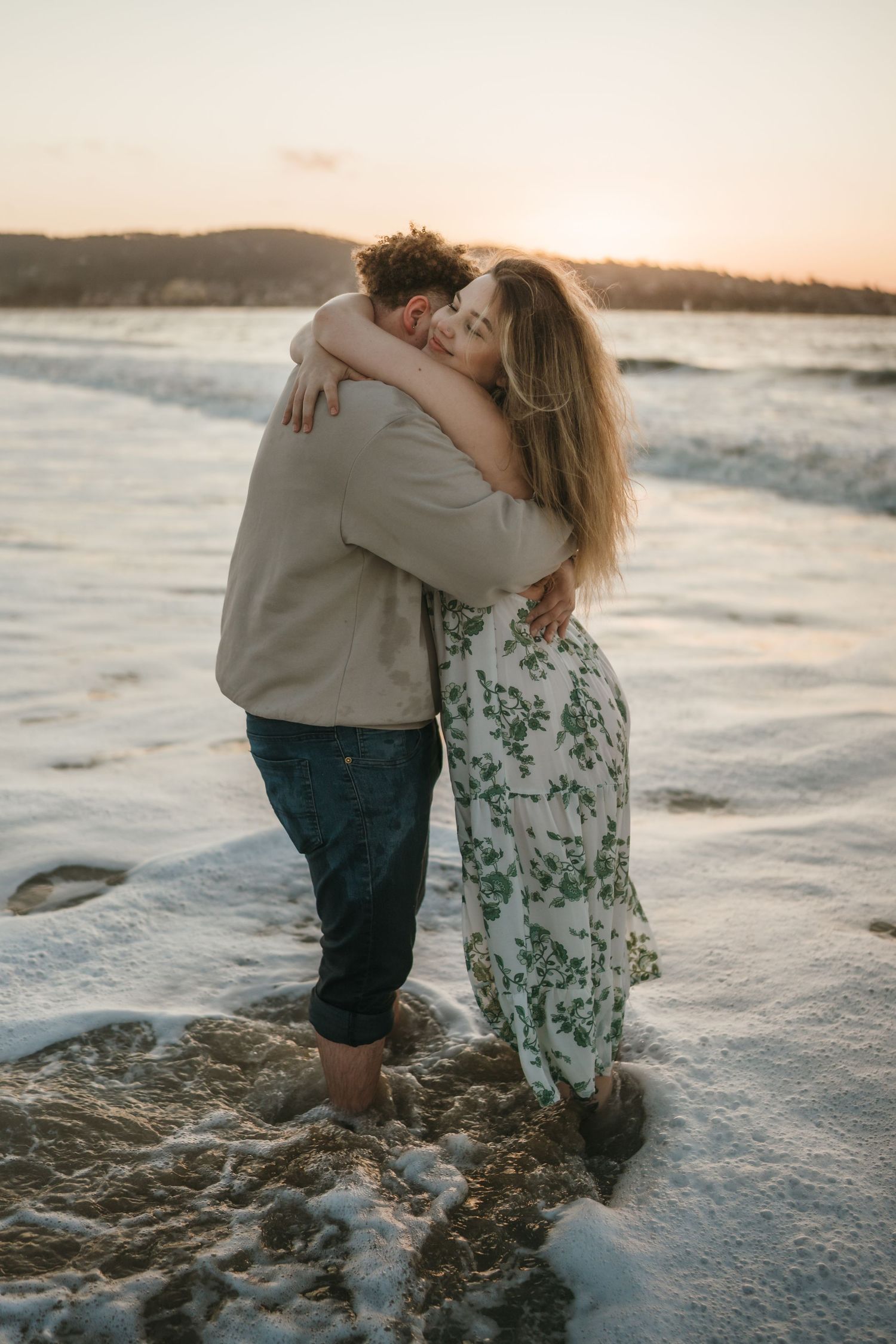 A couple embraces on a sandy beach at sunset while ocean waves roll in behind them.