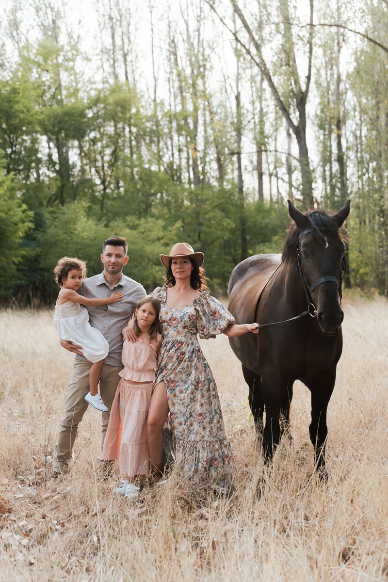 Family in pastel outfits poses together with a dark brown horse in a tall grass field on a sunny day in Hills district Sydney photoshoot