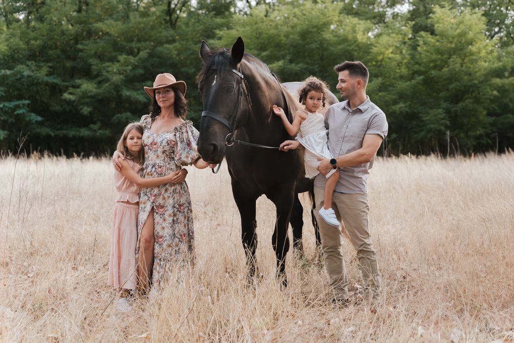 Several people walk together through a tall grass field with sunlight filtering through trees in the background with a horse on a photoshoot