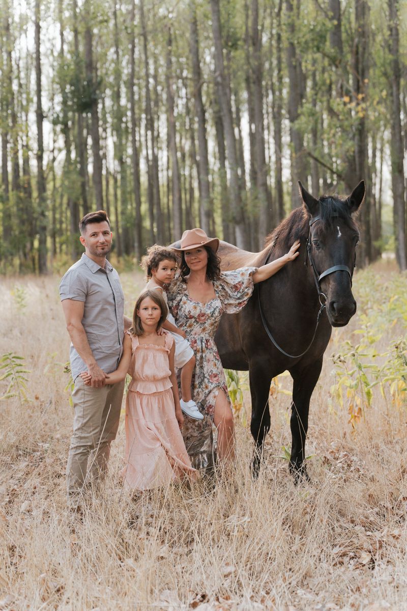 A group poses with a black horse in a sunlit forest meadow filled with tall grass and birch trees.