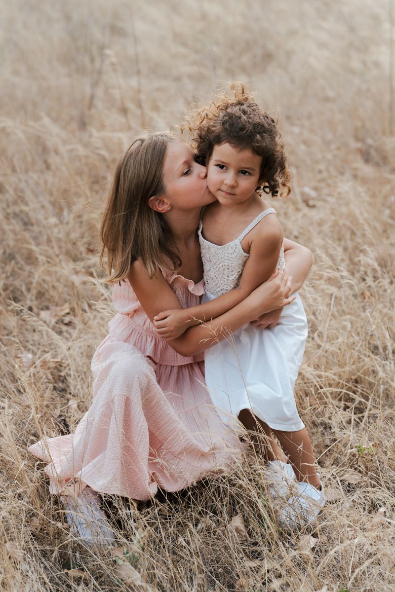 Two girl siblings in pink dresses share moments while sitting in a sunlit field of tall dried grass during a family photoshoot in Sydney