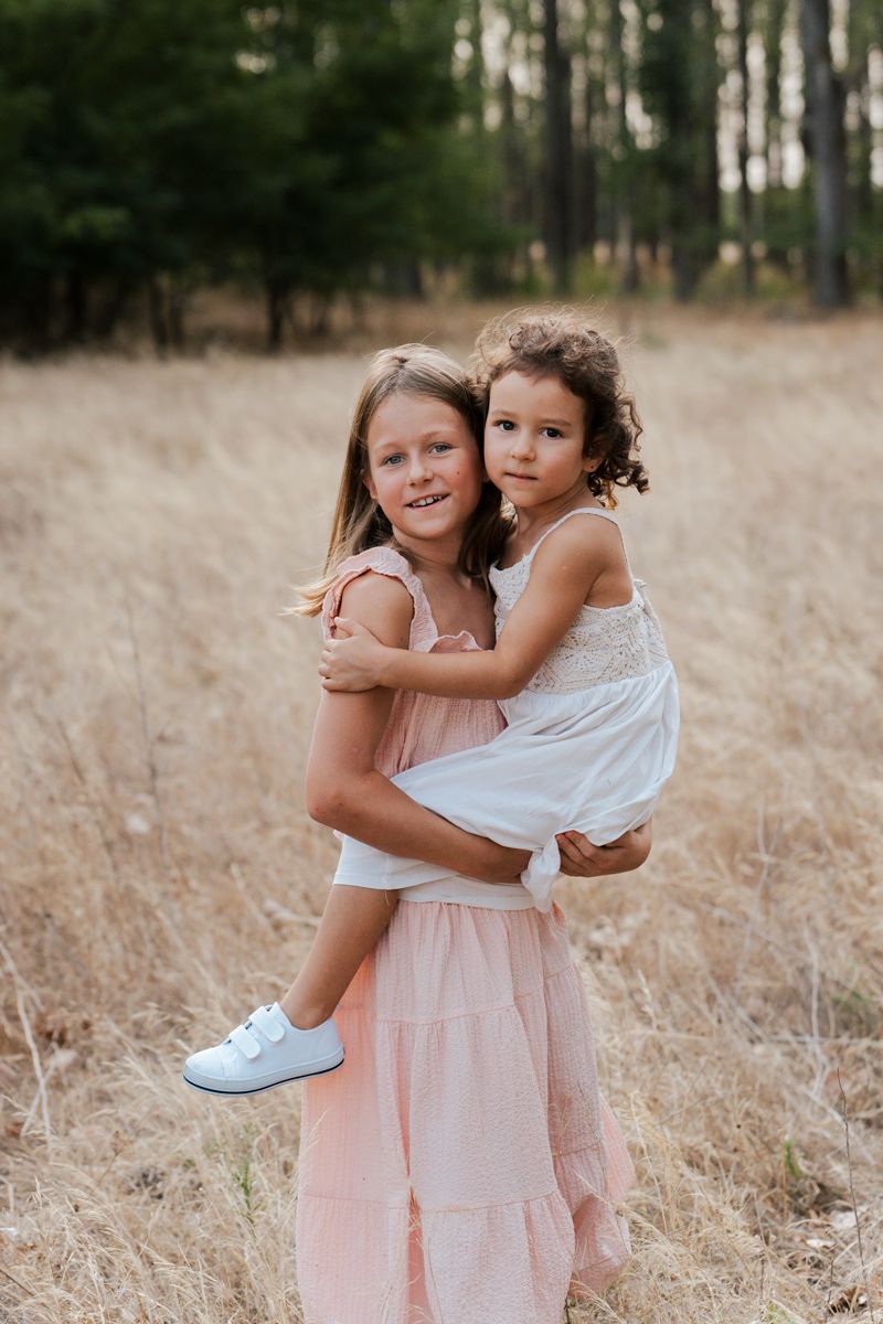 Two children wearing light colored dresses embrace while playing in a golden field at sunset during a family photoshoot in Hills district