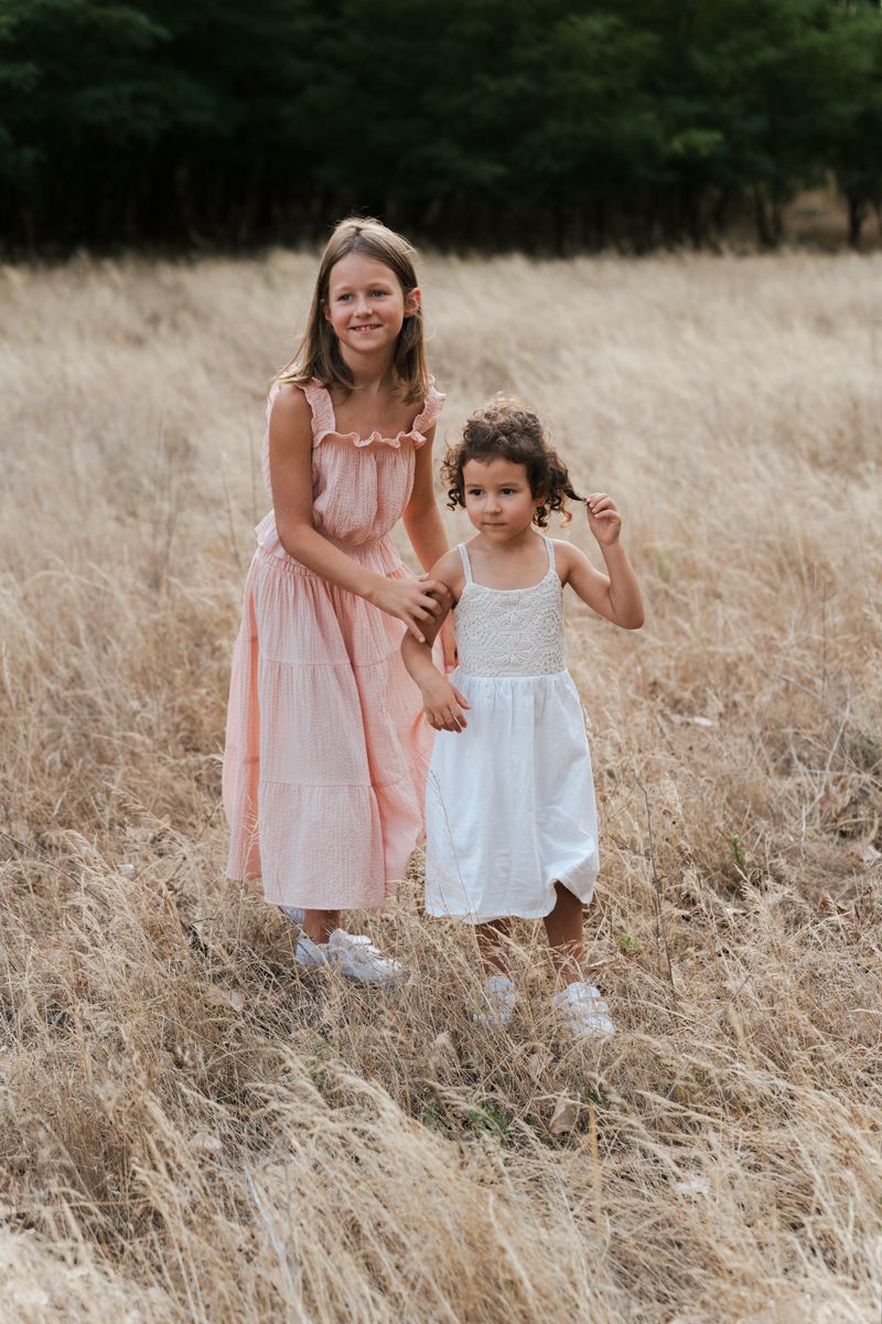 Two siblings in light-colored dresses stand together in a golden wheat field during sunset family photoshoot Hill district