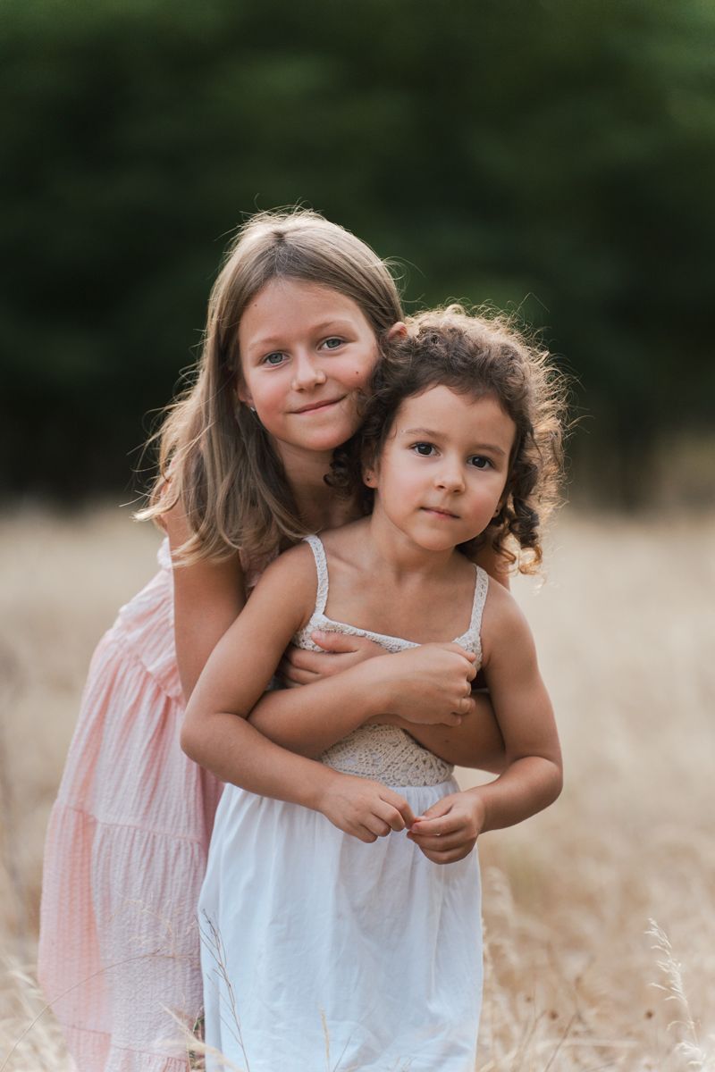 Two children in casual dresses share a sweet embrace while standing in a golden wheat field on a sunny day during a family photoshoot Sydney