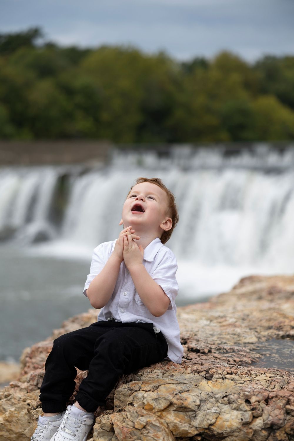 Young child sitting on rocks expressing joy with clasped hands near a cascading waterfall.