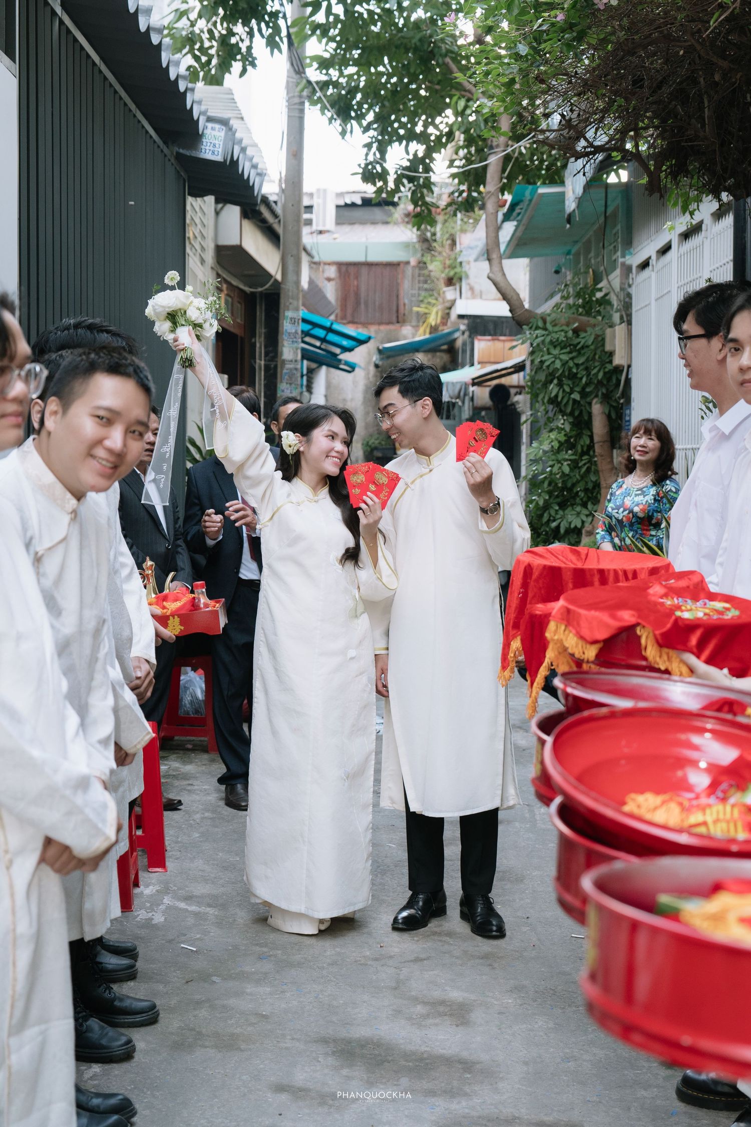 Vietnamese wedding ceremony with couple in white ao dai attire walking through an alley with red gift trays.