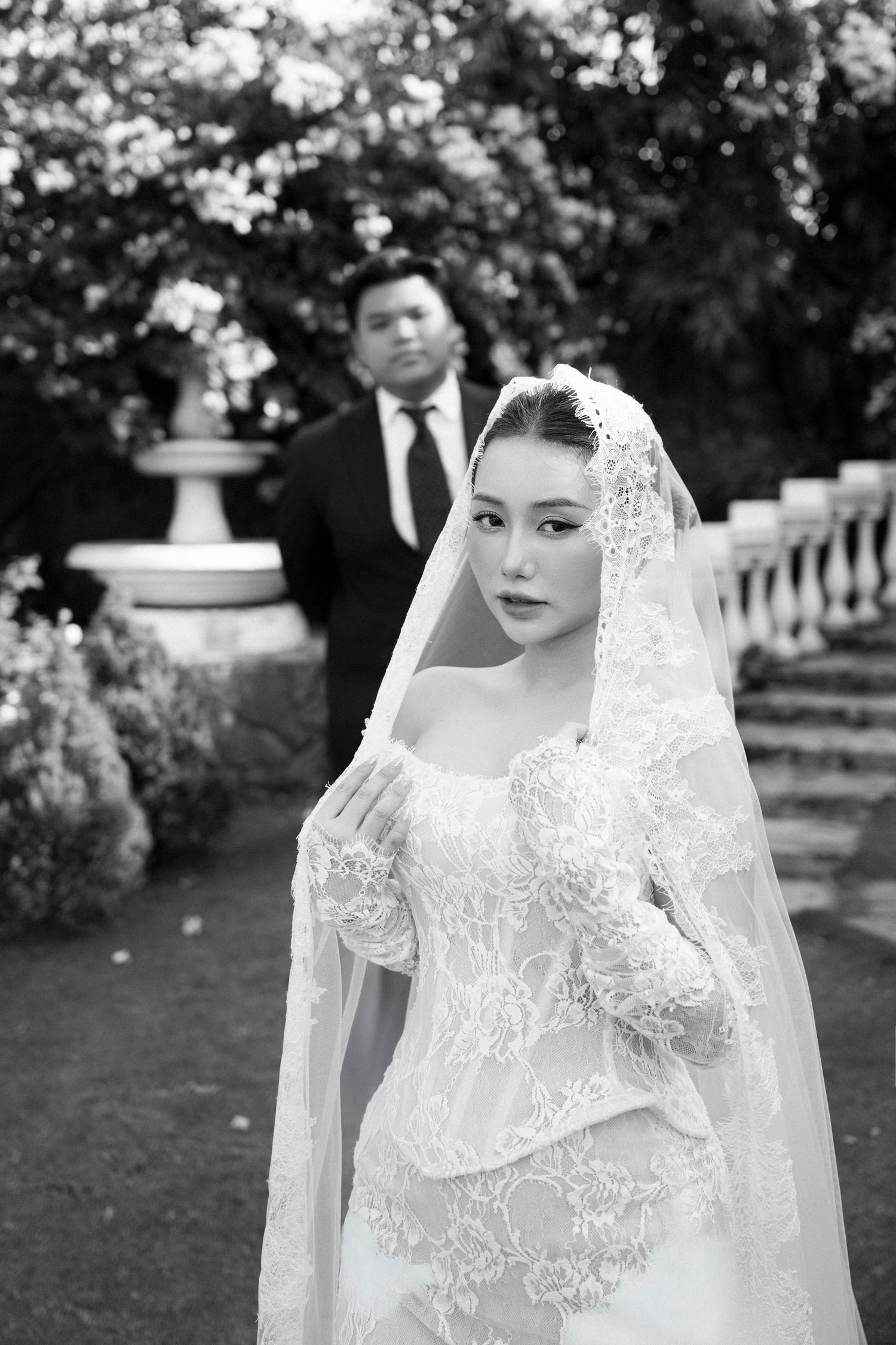 Black and white wedding portrait of bride in lace gown and veil with groom standing in background in a garden.