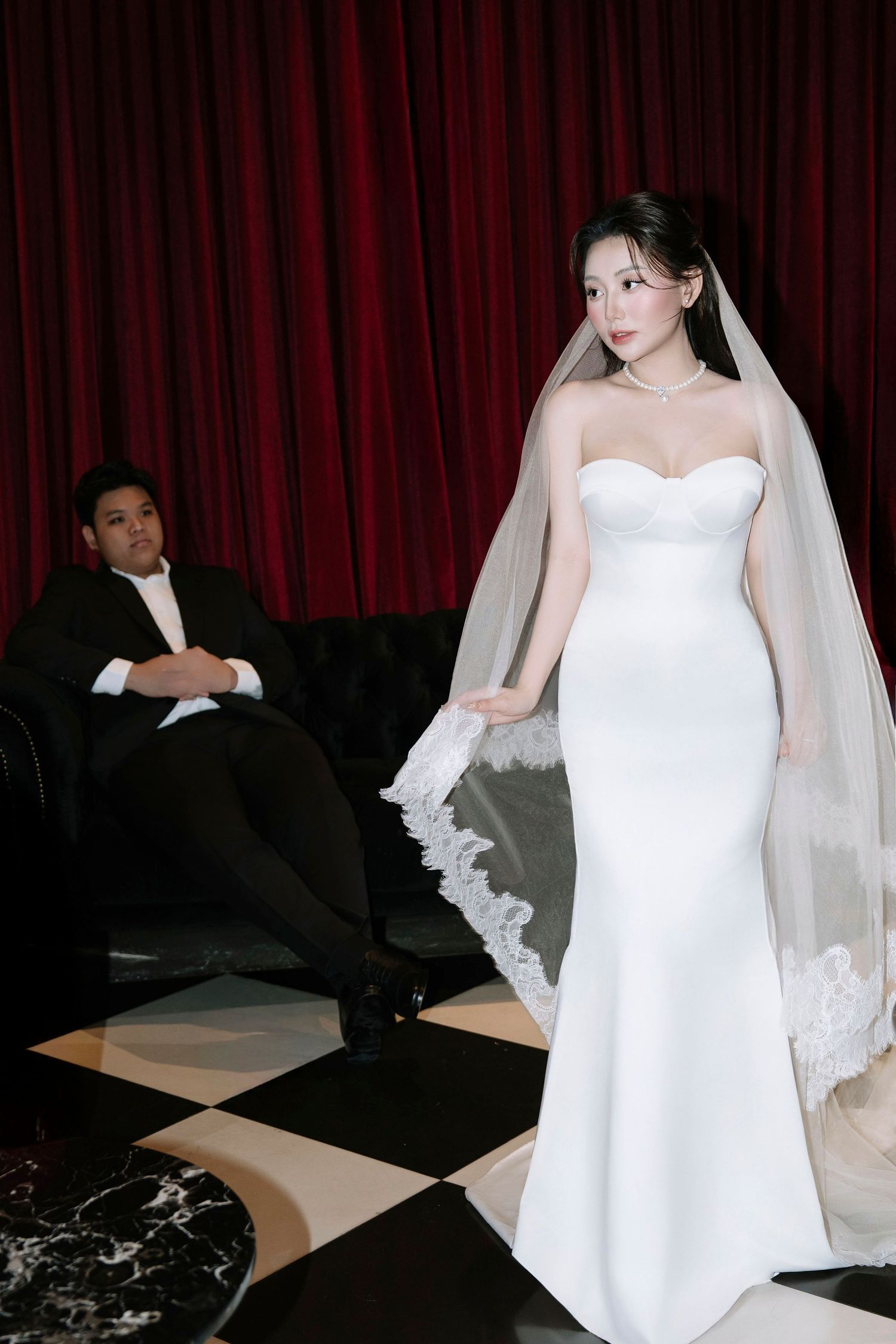 Asian bride in white strapless mermaid gown with lace veil standing before red velvet curtain backdrop.