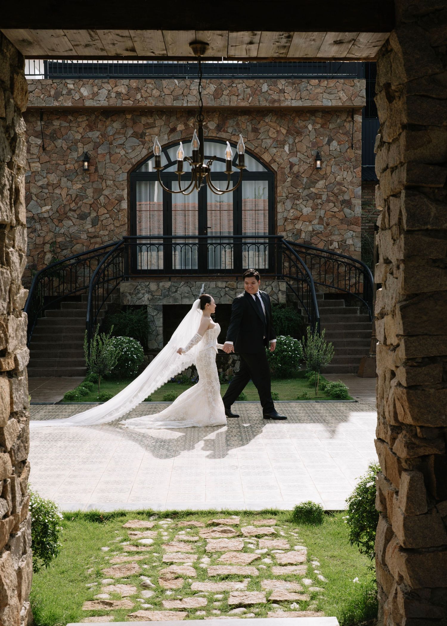 Bride in flowing veil and groom walk hand in hand at stone castle venue.