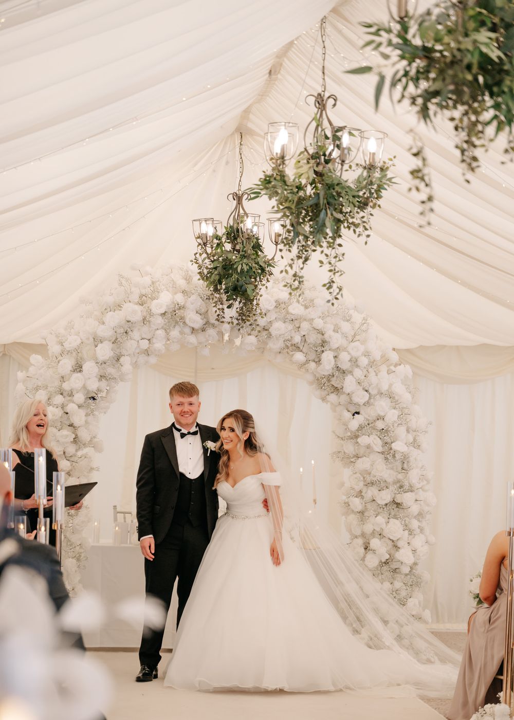 Elegant wedding ceremony setup in white tent with floral arch and candlelit decor.