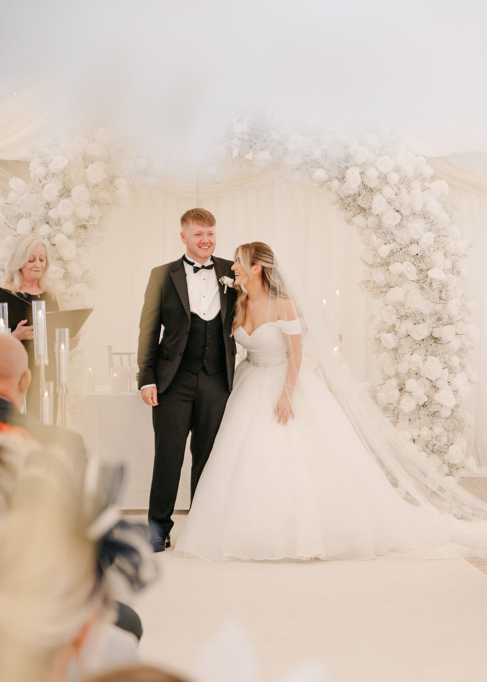 Wedding couple walking together under floral arch in white draped tent.