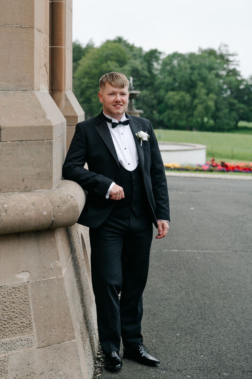 A groom in a classic black tuxedo and bowtie poses against a stone column at an elegant venue.