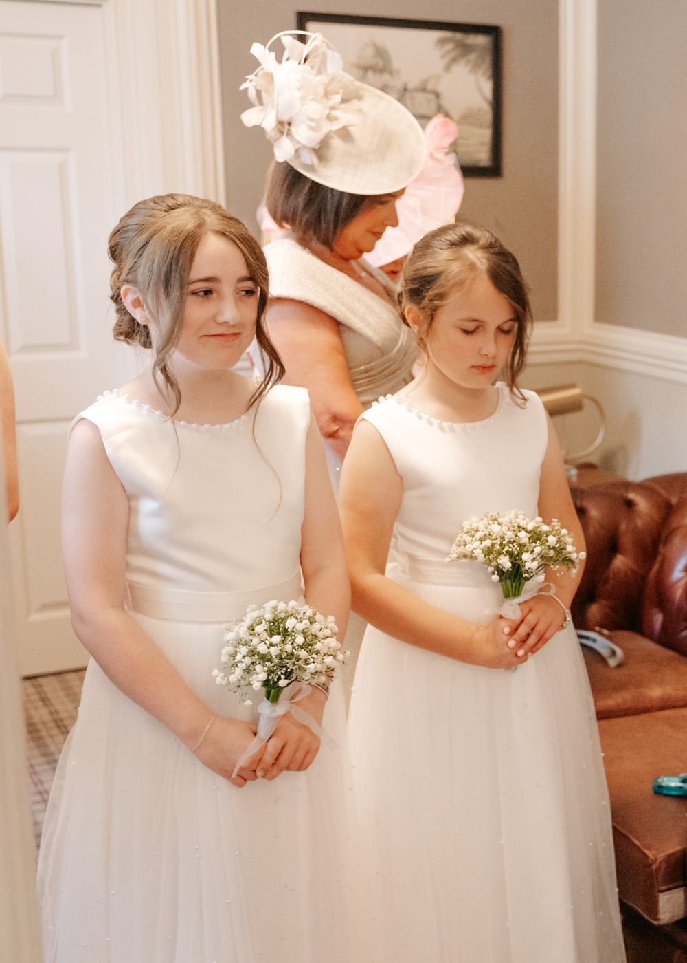 Young bridesmaids in white dresses hold small bouquets while standing in an elegant interior.