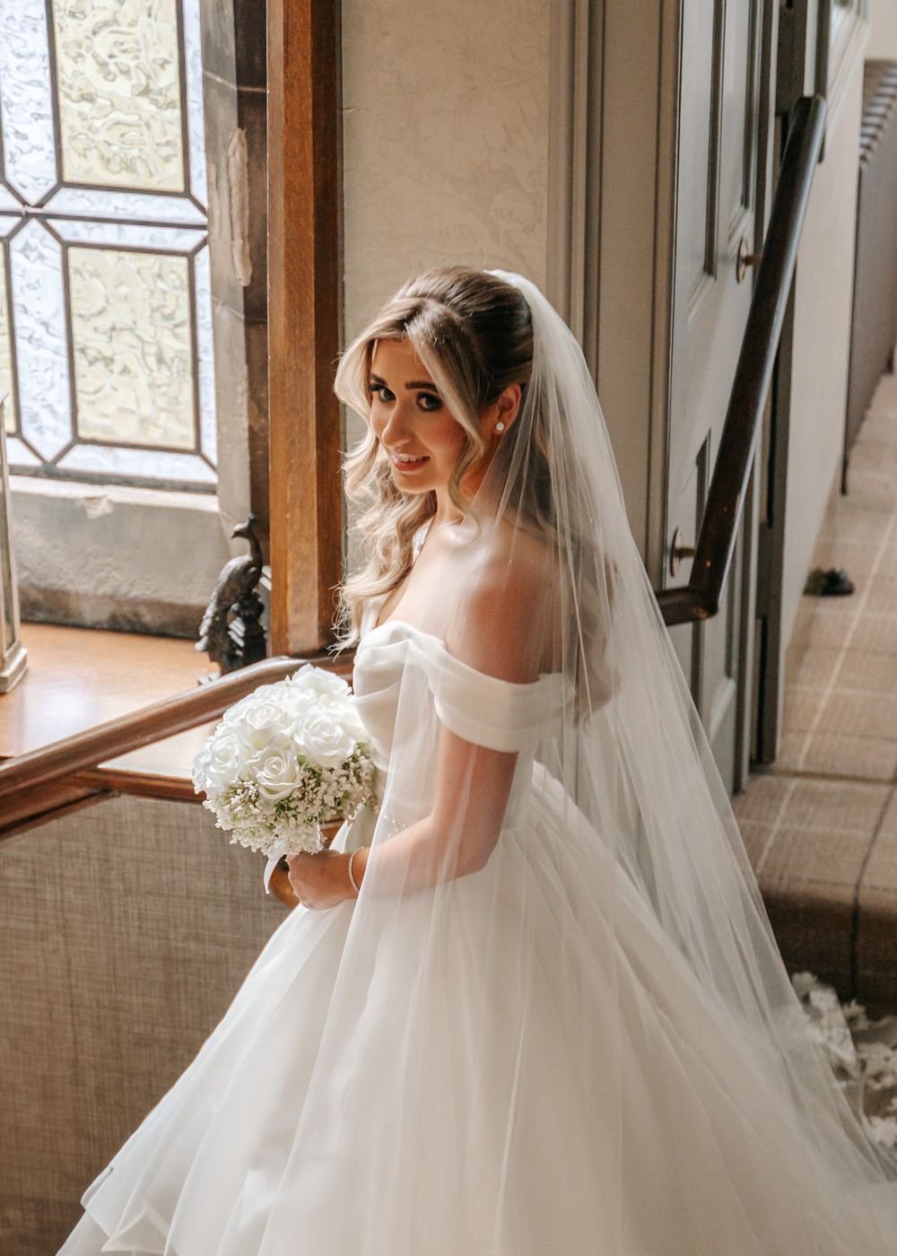 A bride in an off-shoulder ball gown poses by a window while holding a white bouquet.