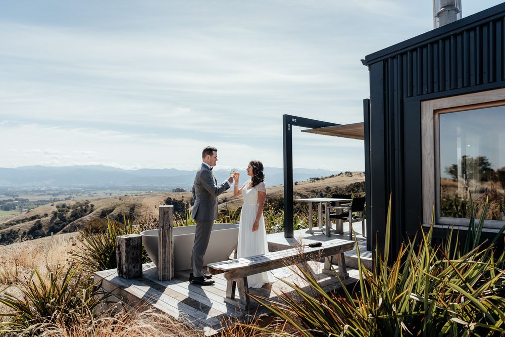 Bride and Groom toast on Whitimanuka's deck with panoramic views of rolling hills and Wairarapa Valley on a sunny afternoon by August & Vine