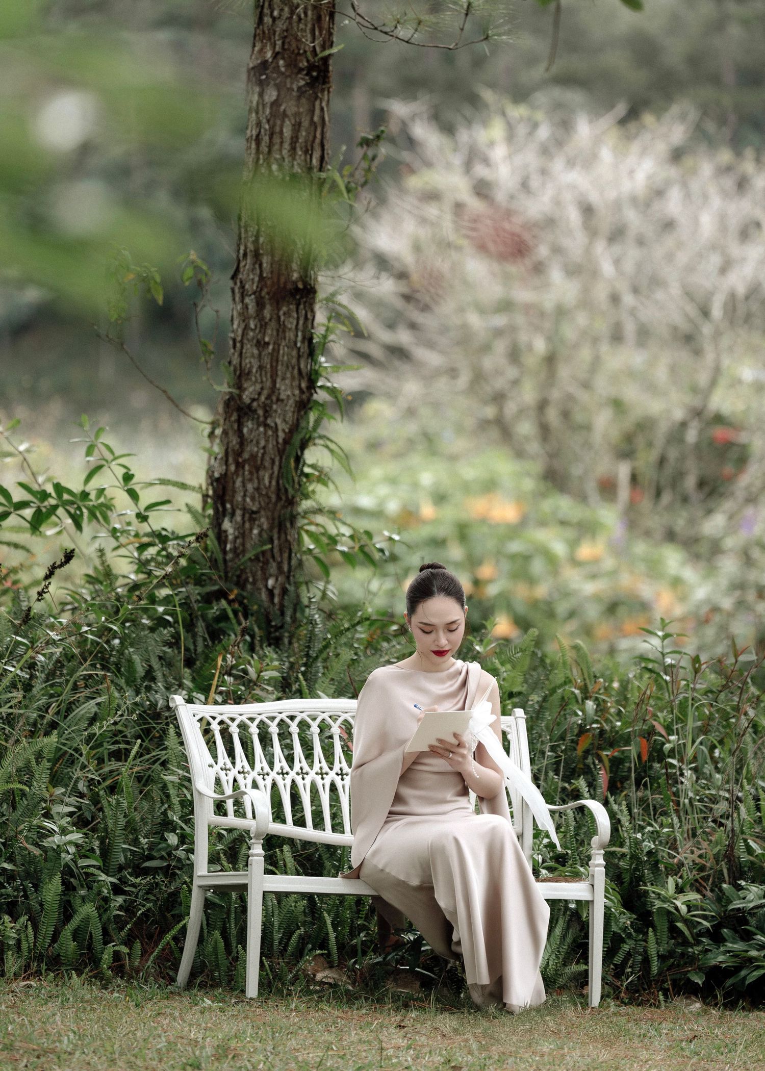 Woman in beige satin dress sitting on white bench in lush garden setting.