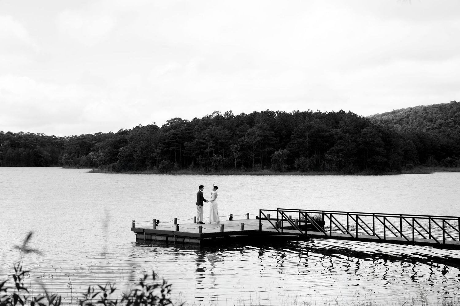 Black and white photo of couple on a wooden dock surrounded by calm lake waters and dense forest treeline.