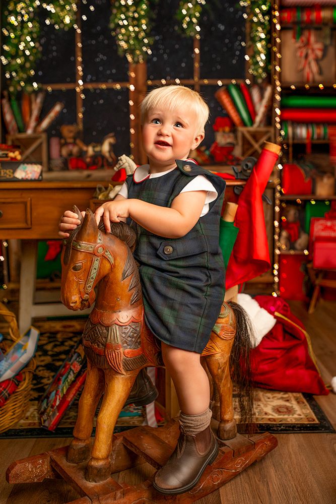 Small child in navy outfit sits on wooden rocking horse with holiday decorations in background.