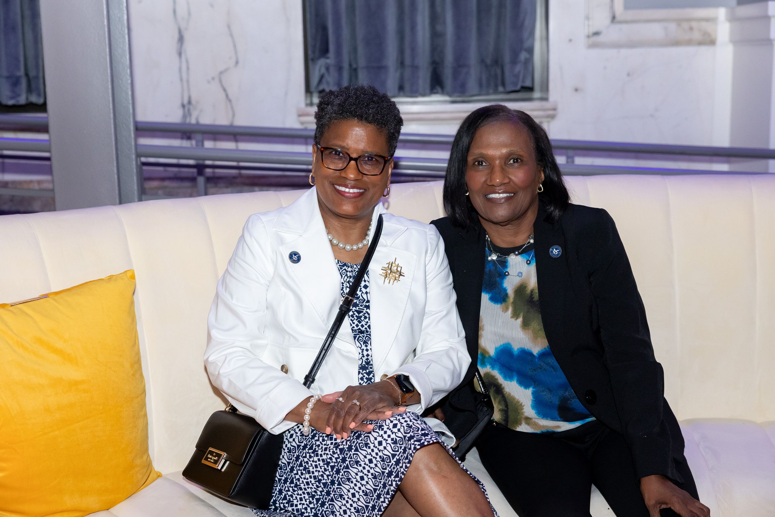 Two women smiling together seated on a white couch at a formal event.