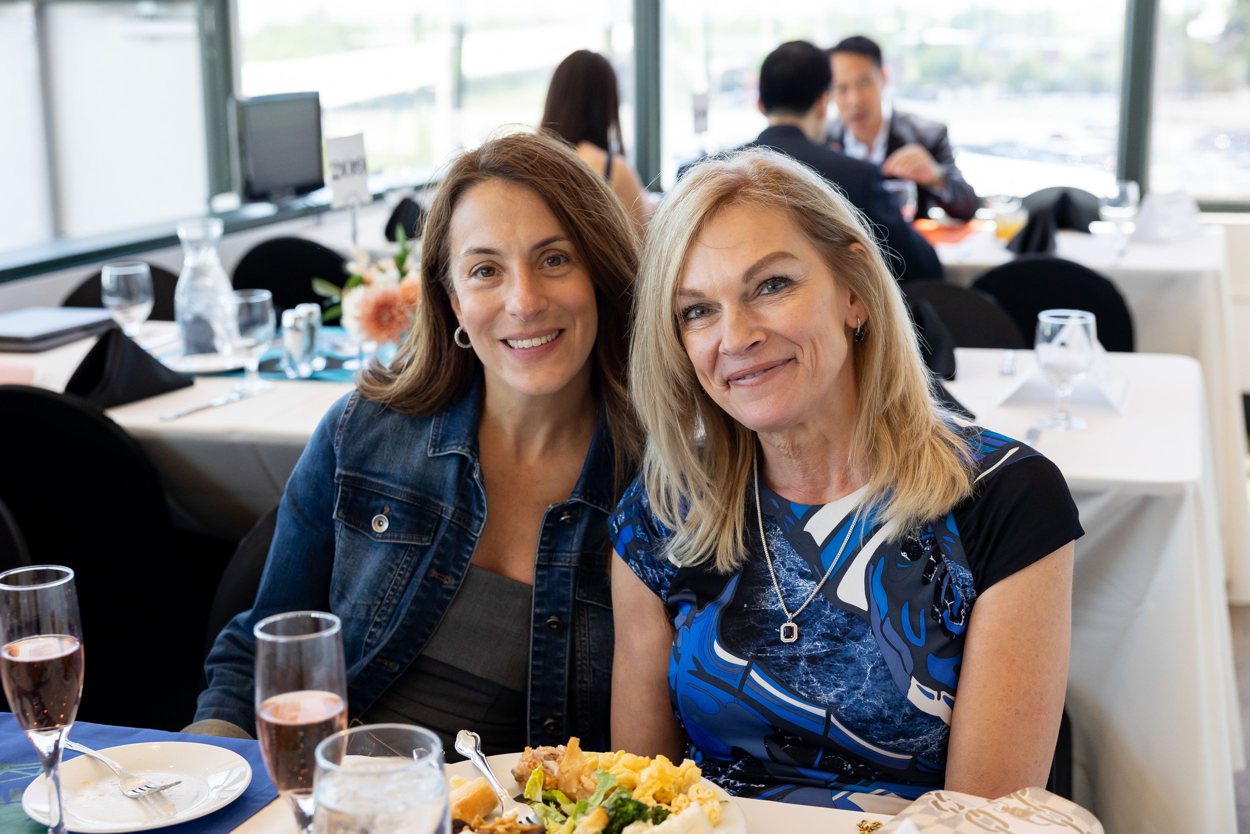 Two smiling women sitting together at a dining table with drinks and food at an indoor event.