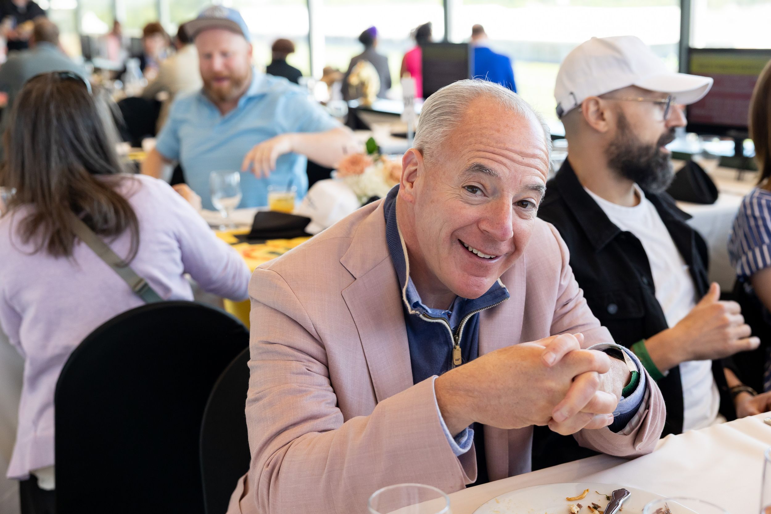Smiling man in a pink blazer sitting at a dining table during a lively indoor event.