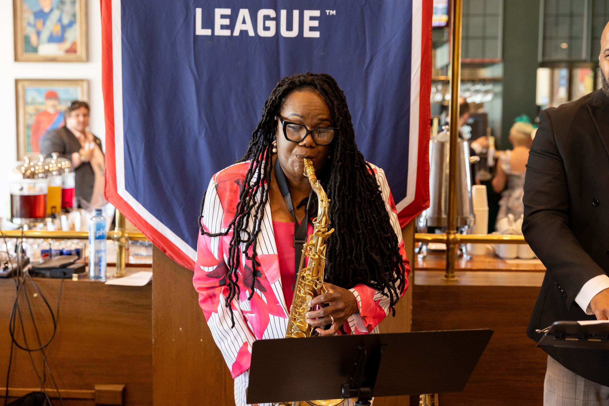 Woman playing saxophone at a League event wearing a pink blazer with long braided hair.