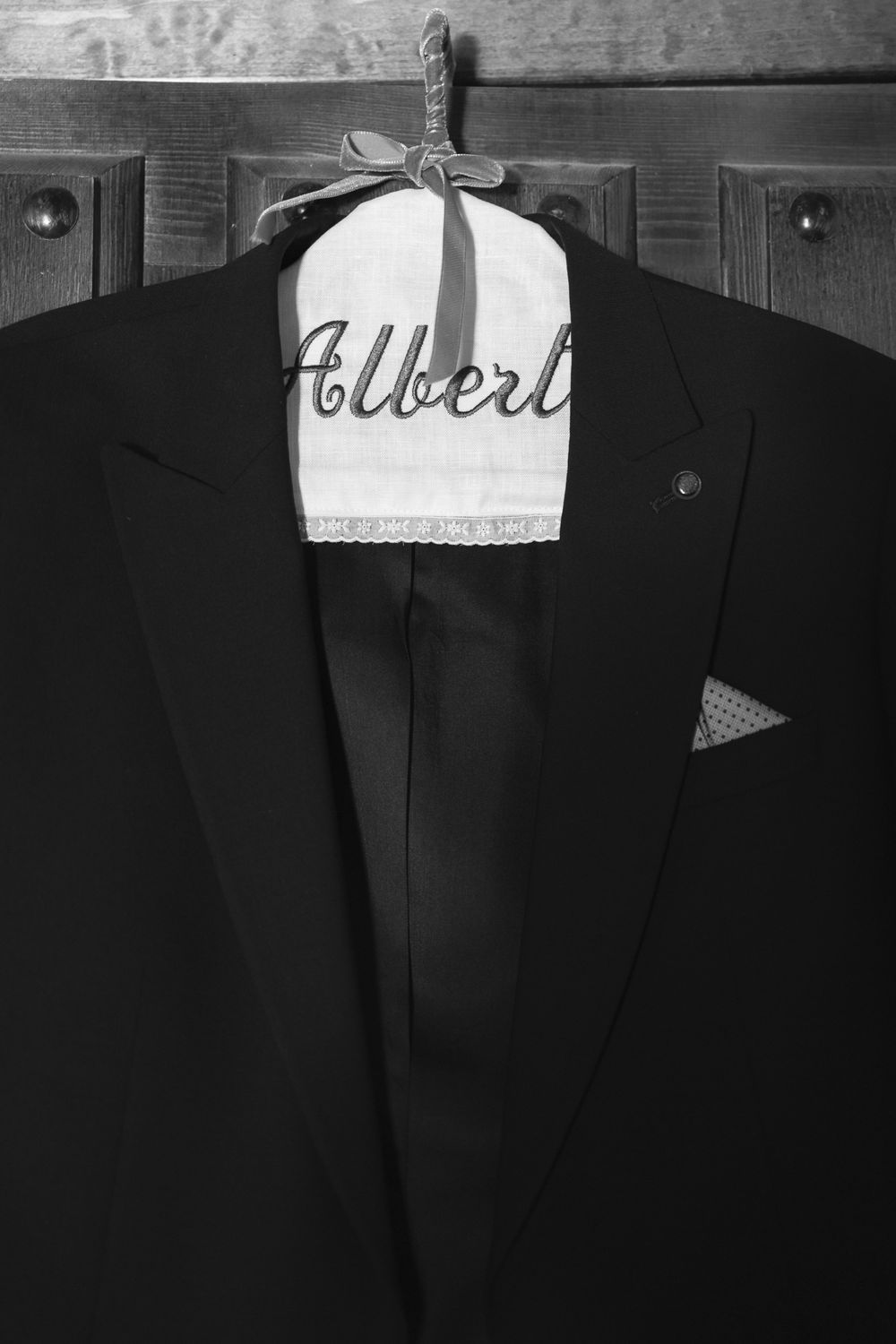 Black formal suit jacket hanging on wooden hanger with embroidered 'Albert' name tag in vintage black and white style.