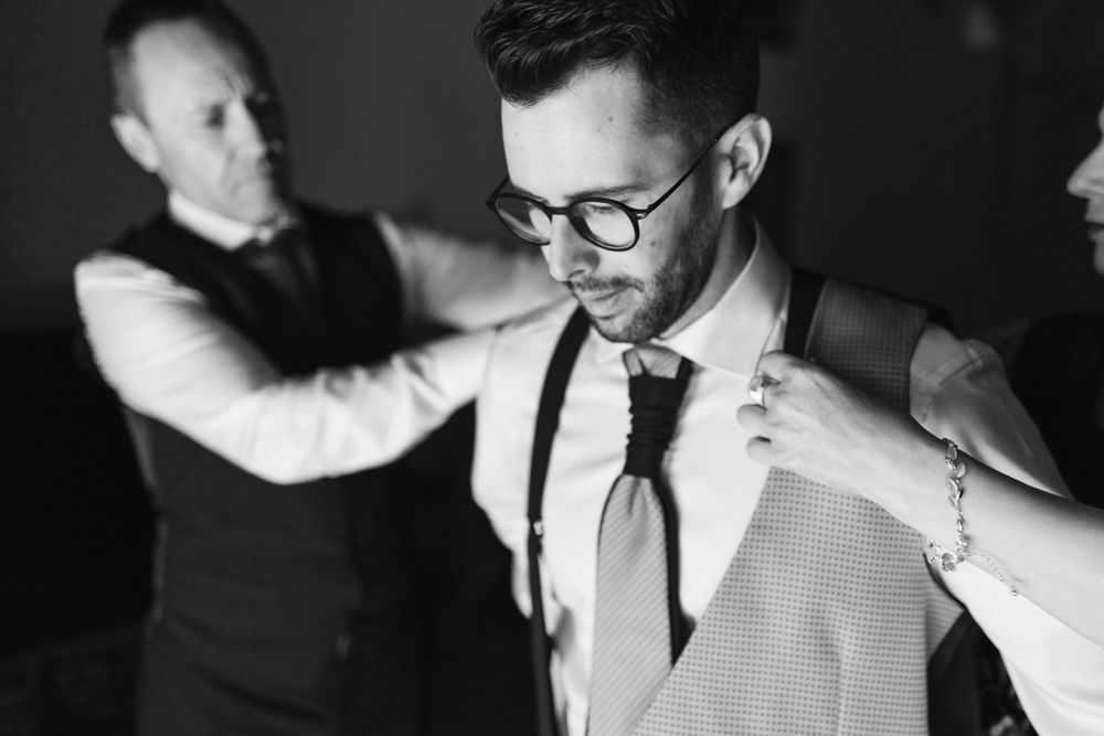 Black and white photograph showing groom in suspenders and tie being helped with formal wear preparations.