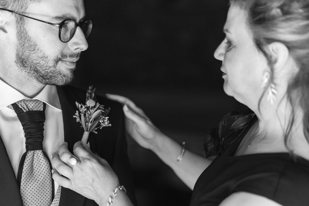 A person adjusts a floral boutonniere on a groom's suit lapel in black and white.