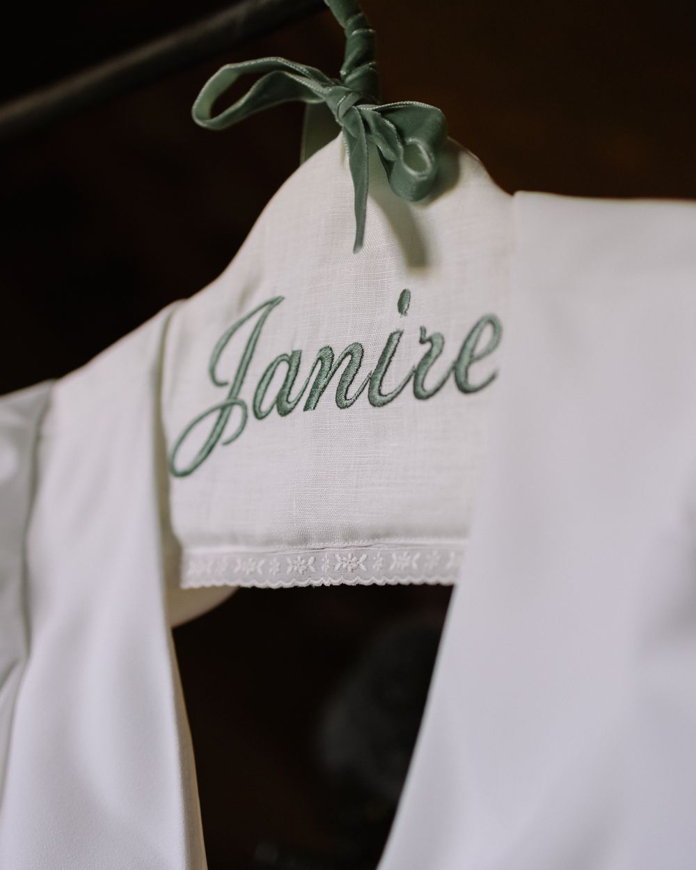 A close-up detail of an embroidered 'Janice' name on a white garment hanger with a sage green ribbon.