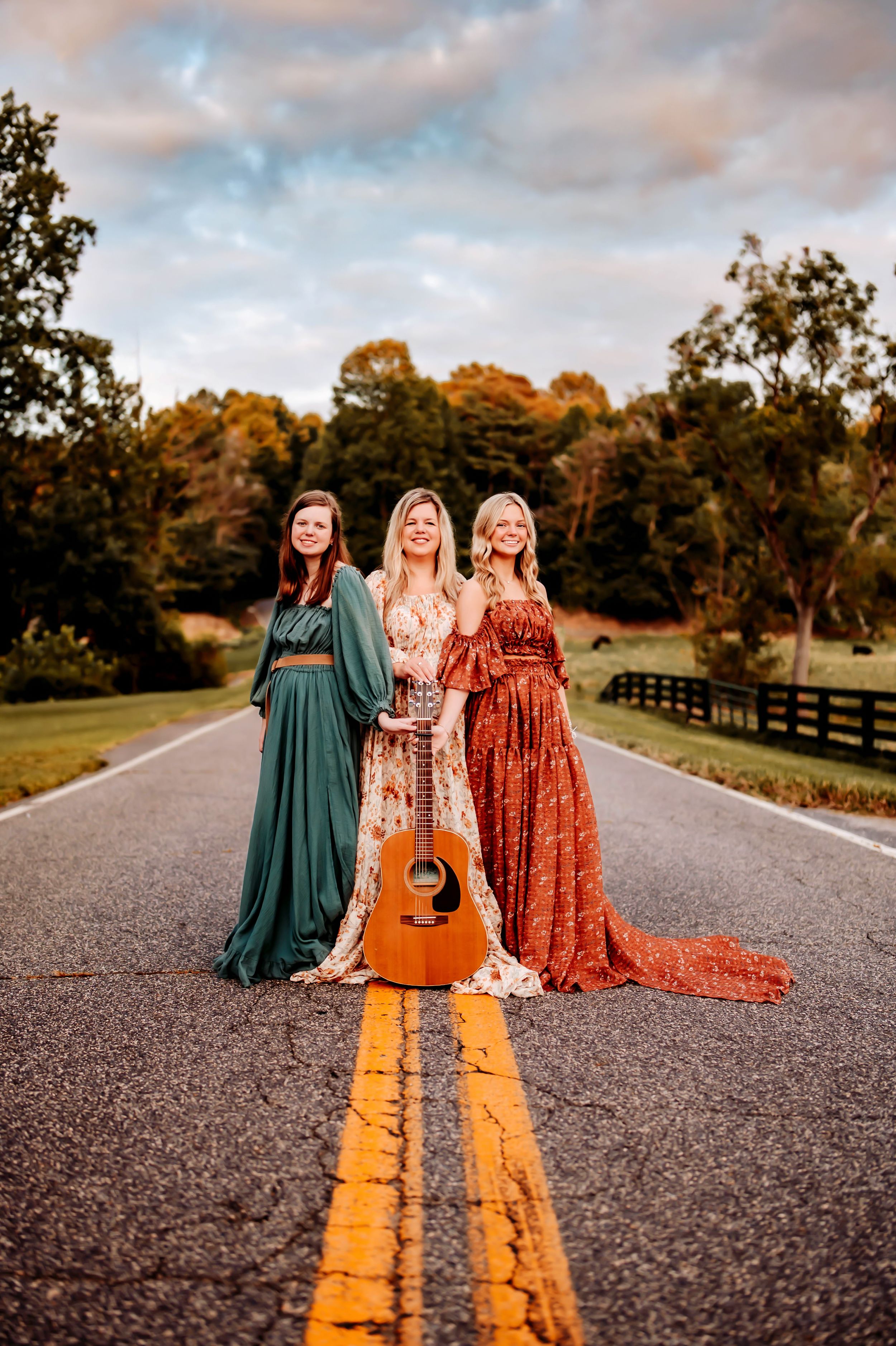 Three people in flowing maxi dresses stand on a country road with an acoustic guitar at sunset, framed by autumn trees.