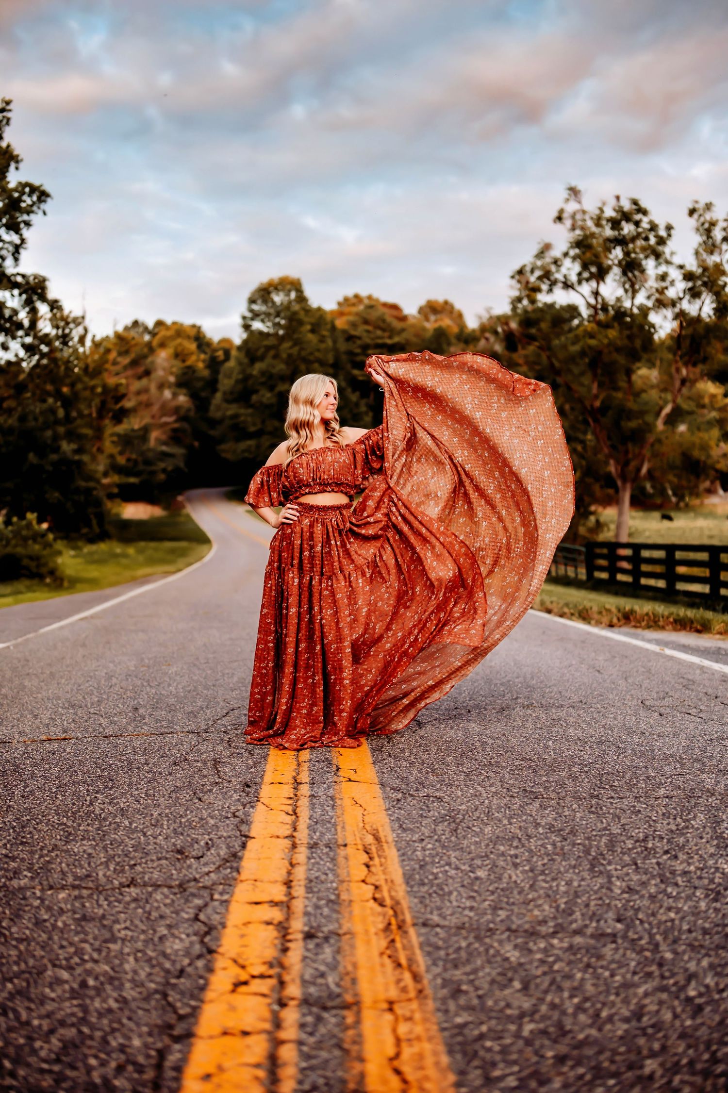 A flowing red dress billows dramatically across a rural road with yellow lines and wooden fences at sunset.