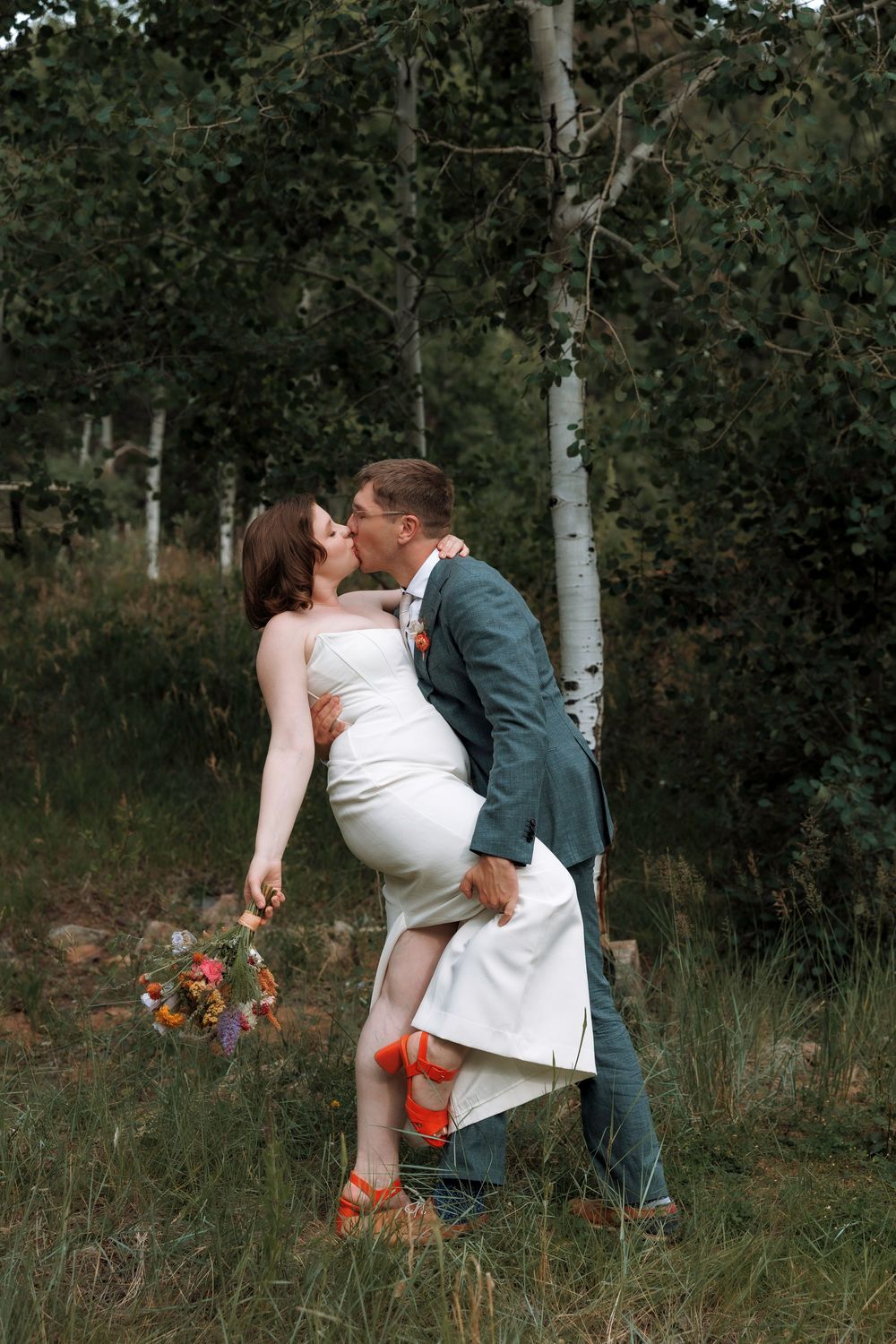 Groom dipping bride for a kiss near birch trees while she holds colorful wildflower bouquet.