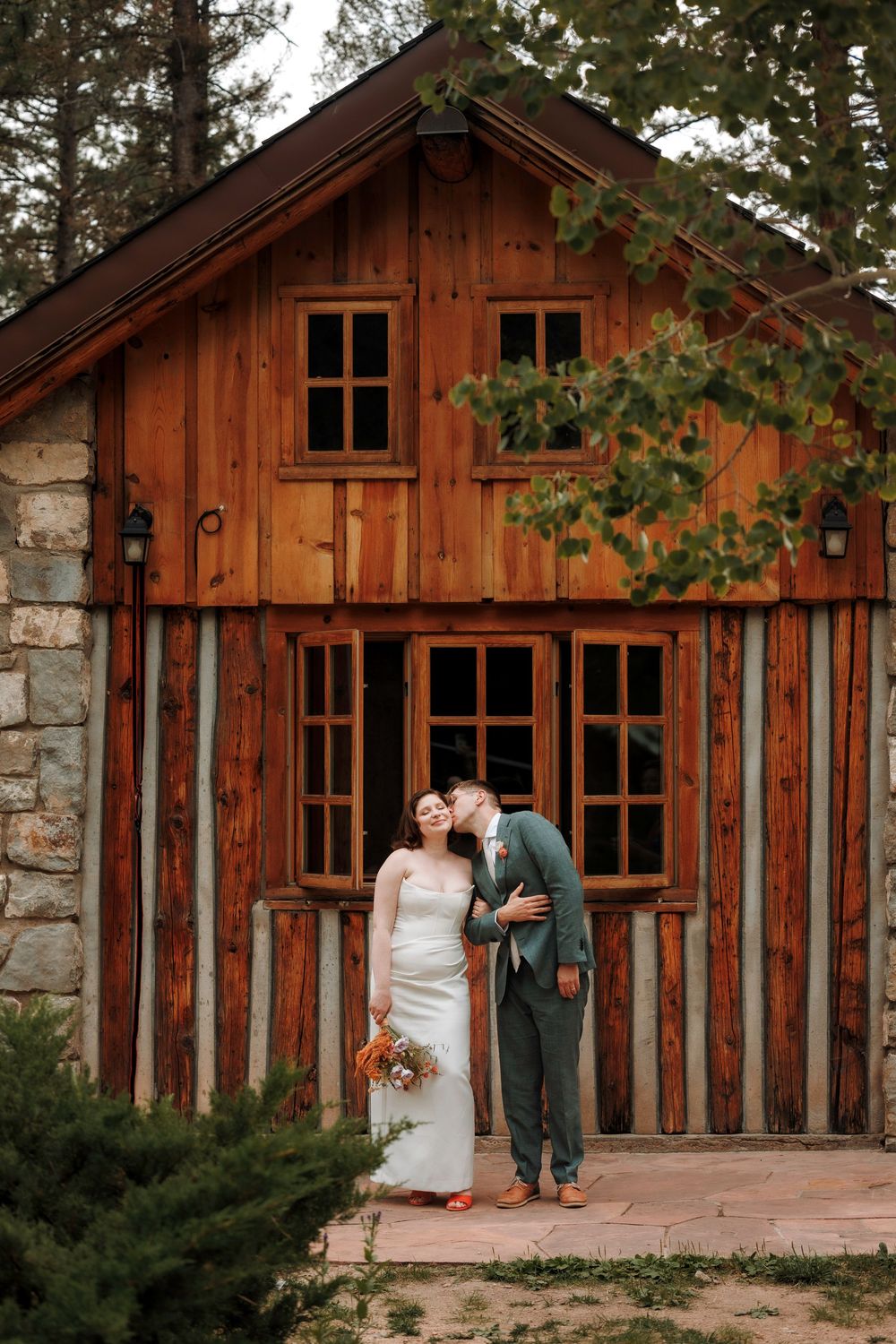 Bride and groom kissing in front of a rustic wooden cabin surrounded by pine trees.