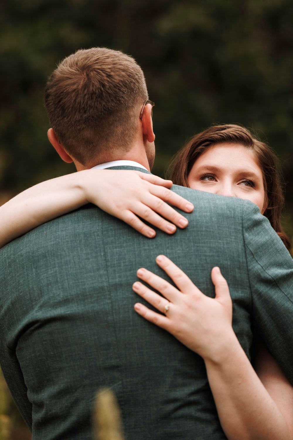 Bride with engagement ring hugging groom in green suit from behind in a forest setting.