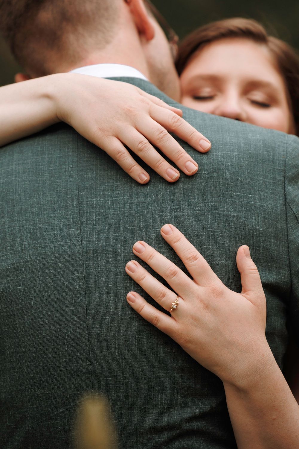 Close-up of bride's hands with engagement ring embracing groom in a gray-green suit.