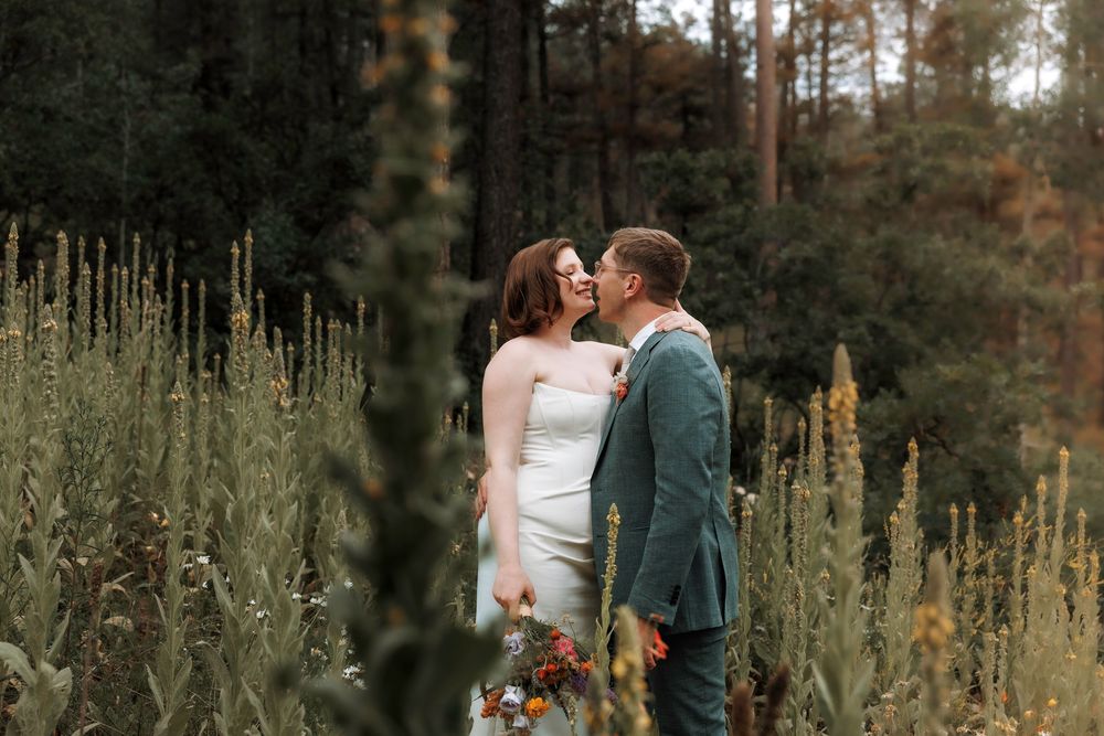 Bride and groom sharing a kiss surrounded by tall wildflowers in a lush forest meadow.