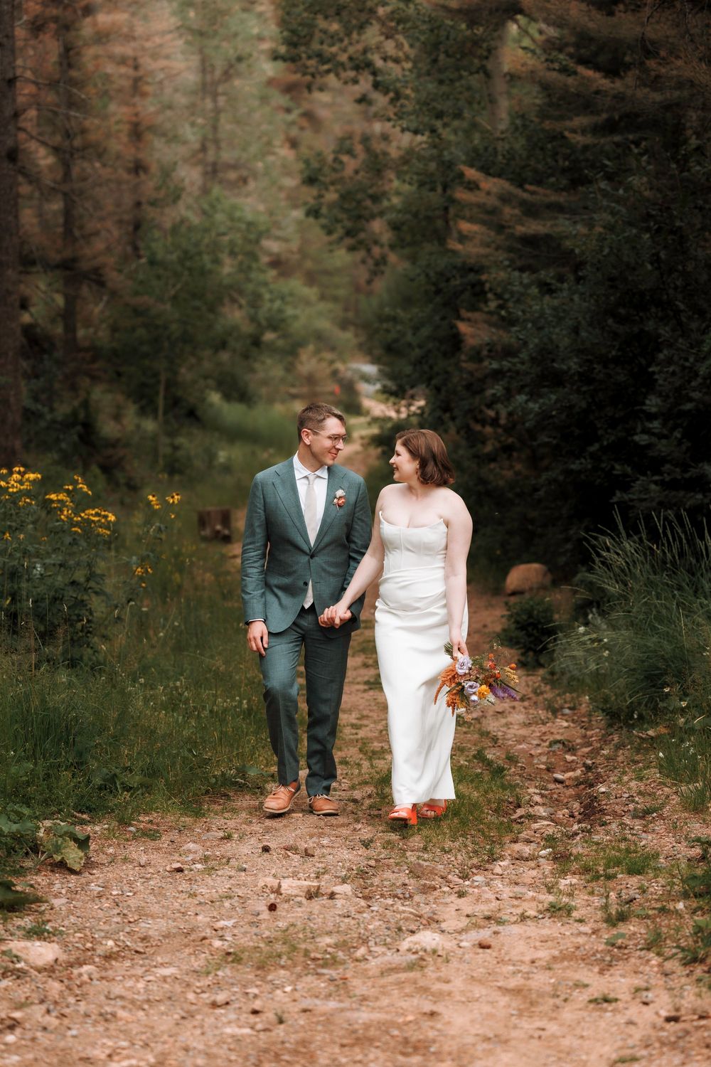Bride and groom holding hands and walking along a scenic forest dirt path together.