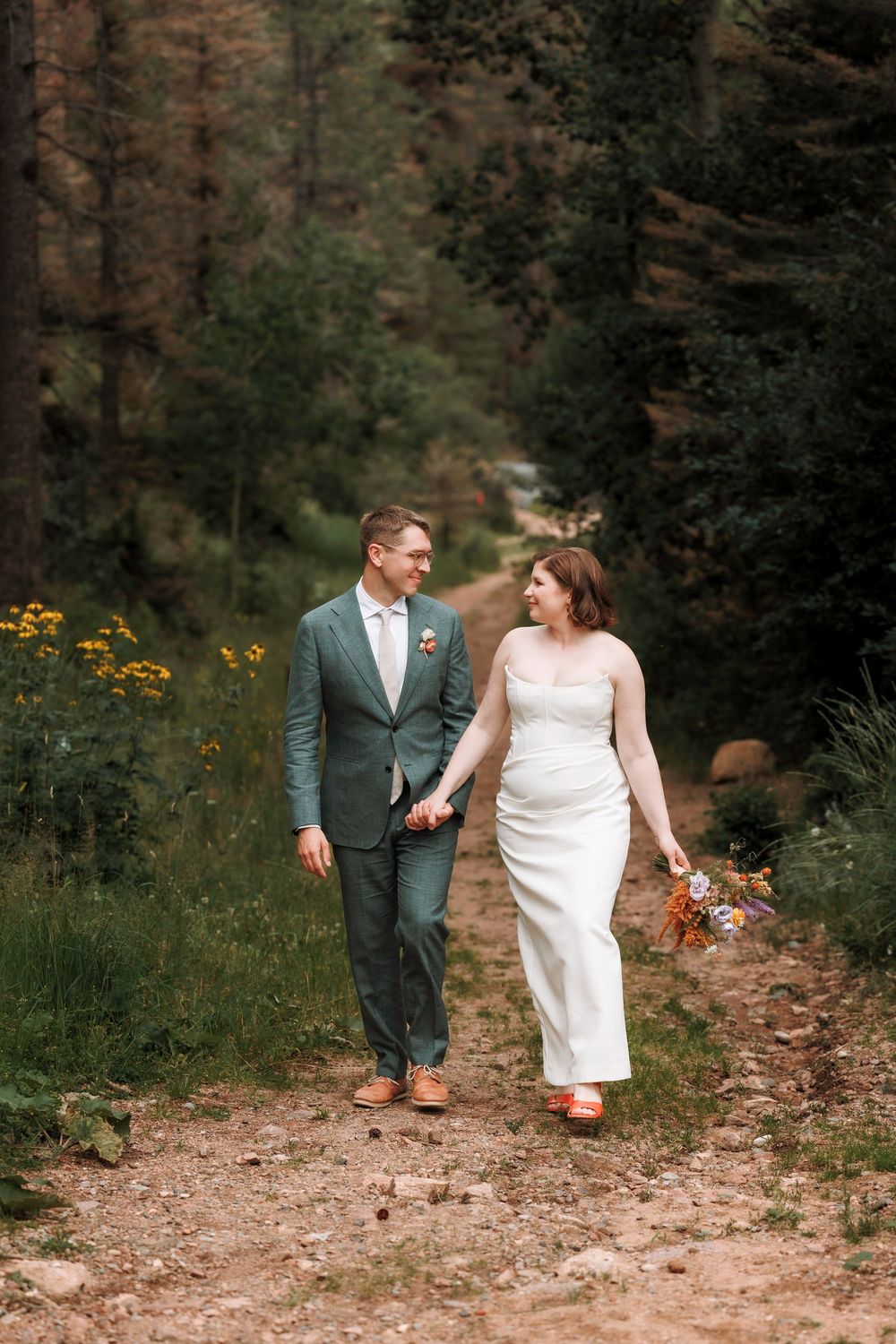 Smiling bride and groom walking hand in hand on a forest trail with wildflowers nearby.