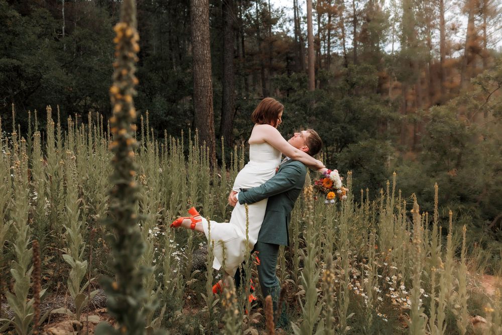 Groom lifting bride in white dress among tall wildflowers in a lush green forest meadow.