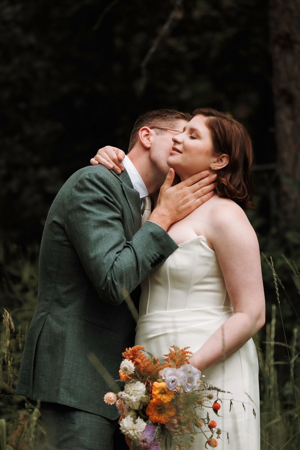 Groom tenderly kissing bride while holding her face as she holds a colorful bridal bouquet.