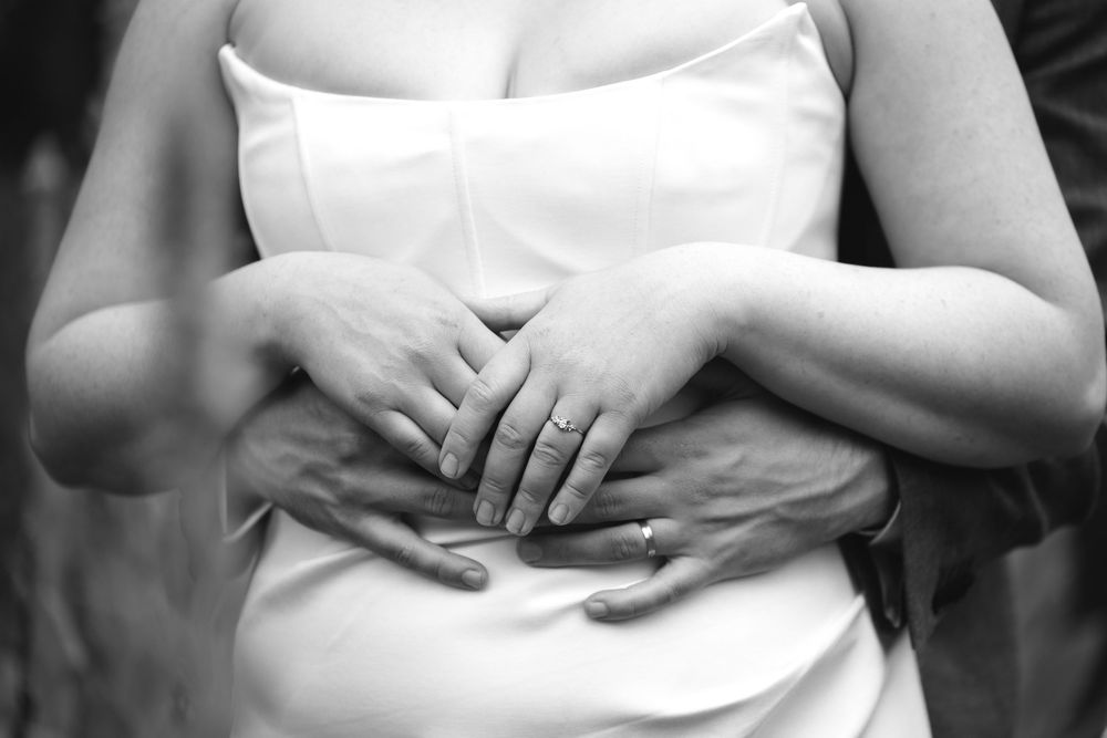 Close-up black and white photo of bride and groom hands with wedding rings embracing at waist.