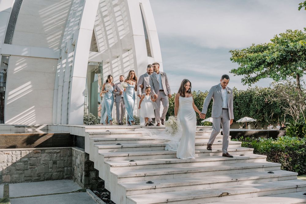 Wedding party members walk down grand white stone steps in a cascading line at an elegant outdoor venue.