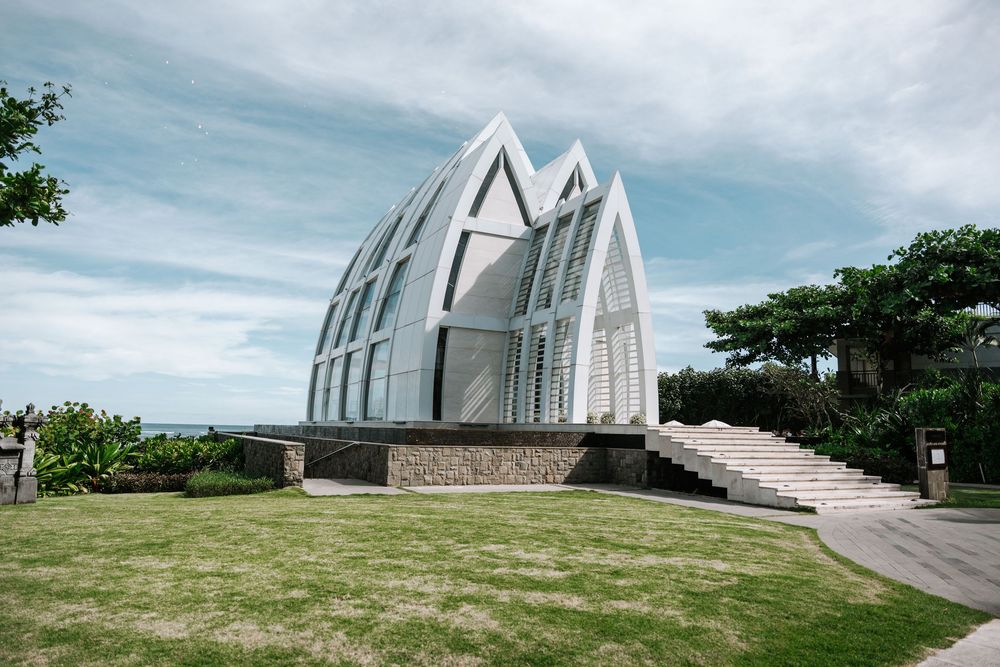 Modern white glass and steel wedding chapel with geometric architecture sits on a hilltop overlooking the ocean landscape.