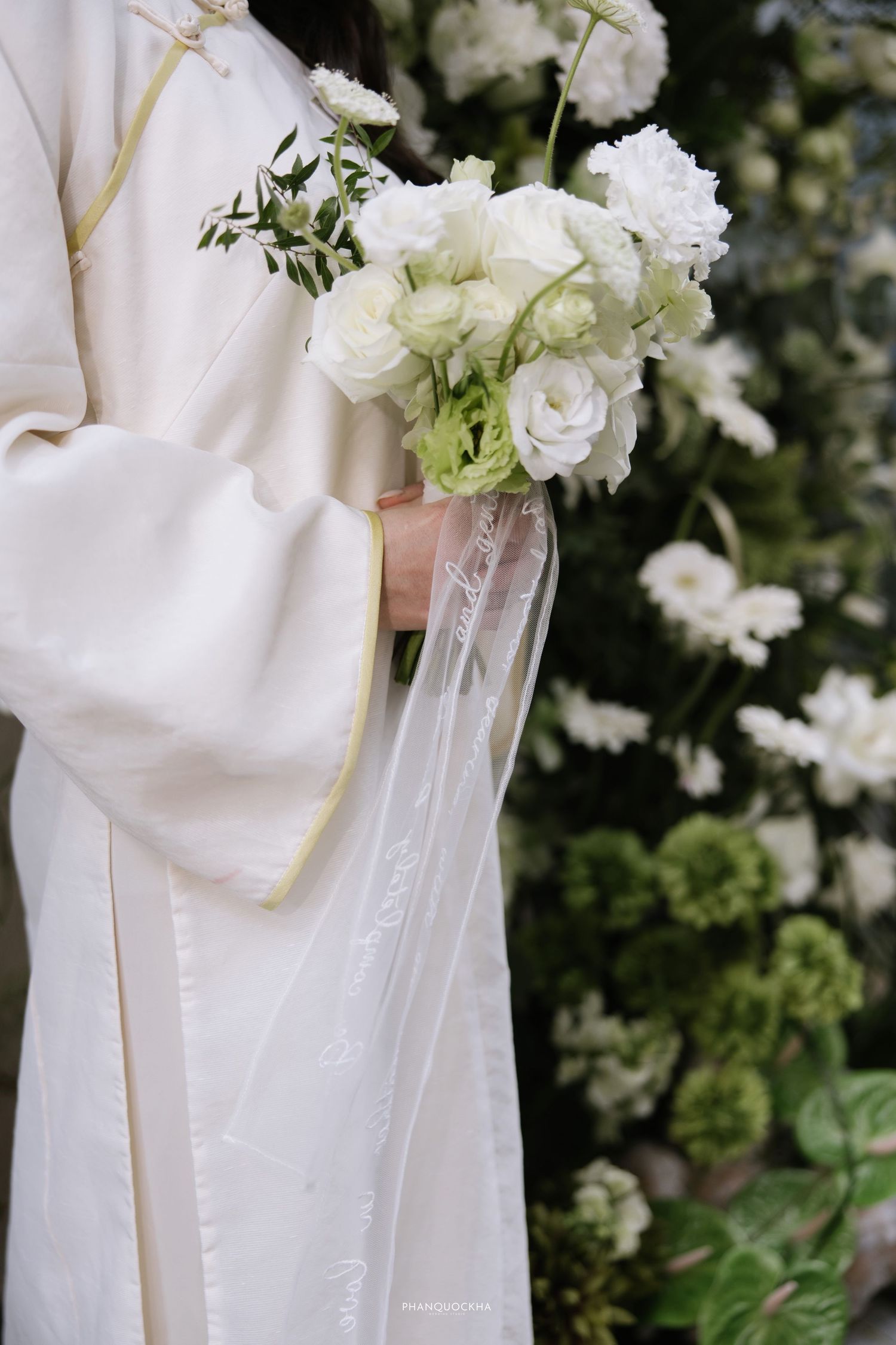 Close-up of bride in white ao dai holding white rose bouquet with sheer ribbon.