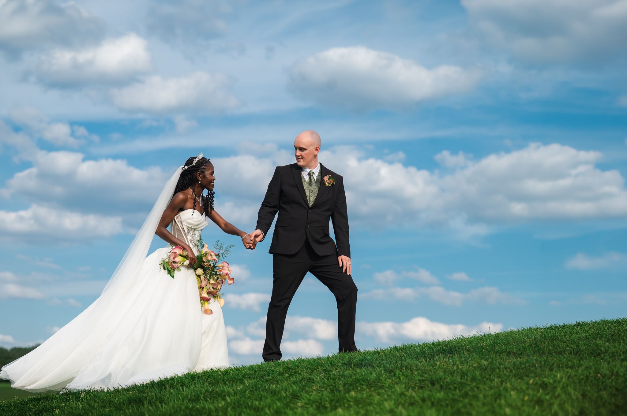 Newlyweds standing atop a grassy hill against a bright blue sky with white clouds.